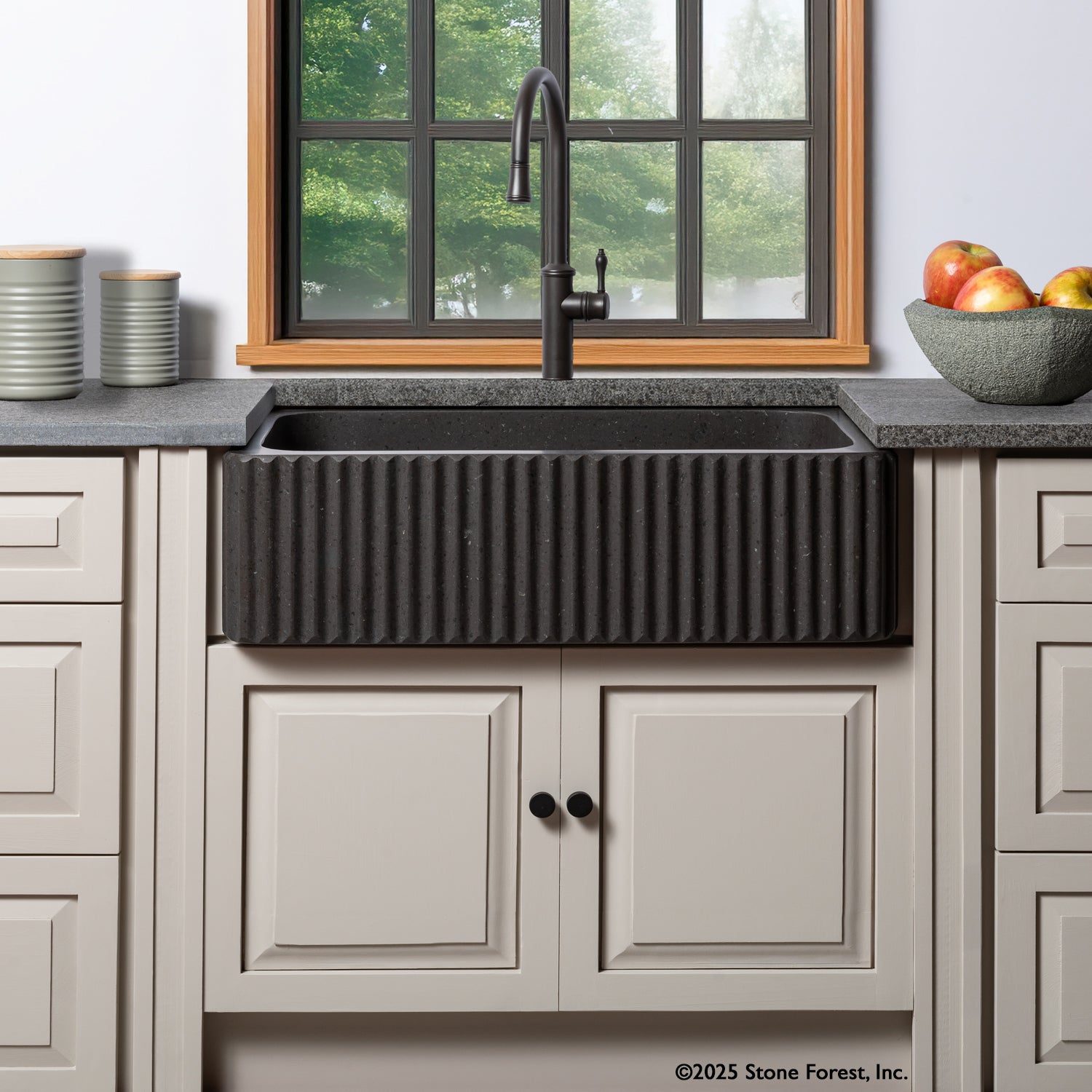 A fluted front farmhouse sink made of noce basalt, installed in a kitchen setting with a black faucet and light-colored cabinetry.