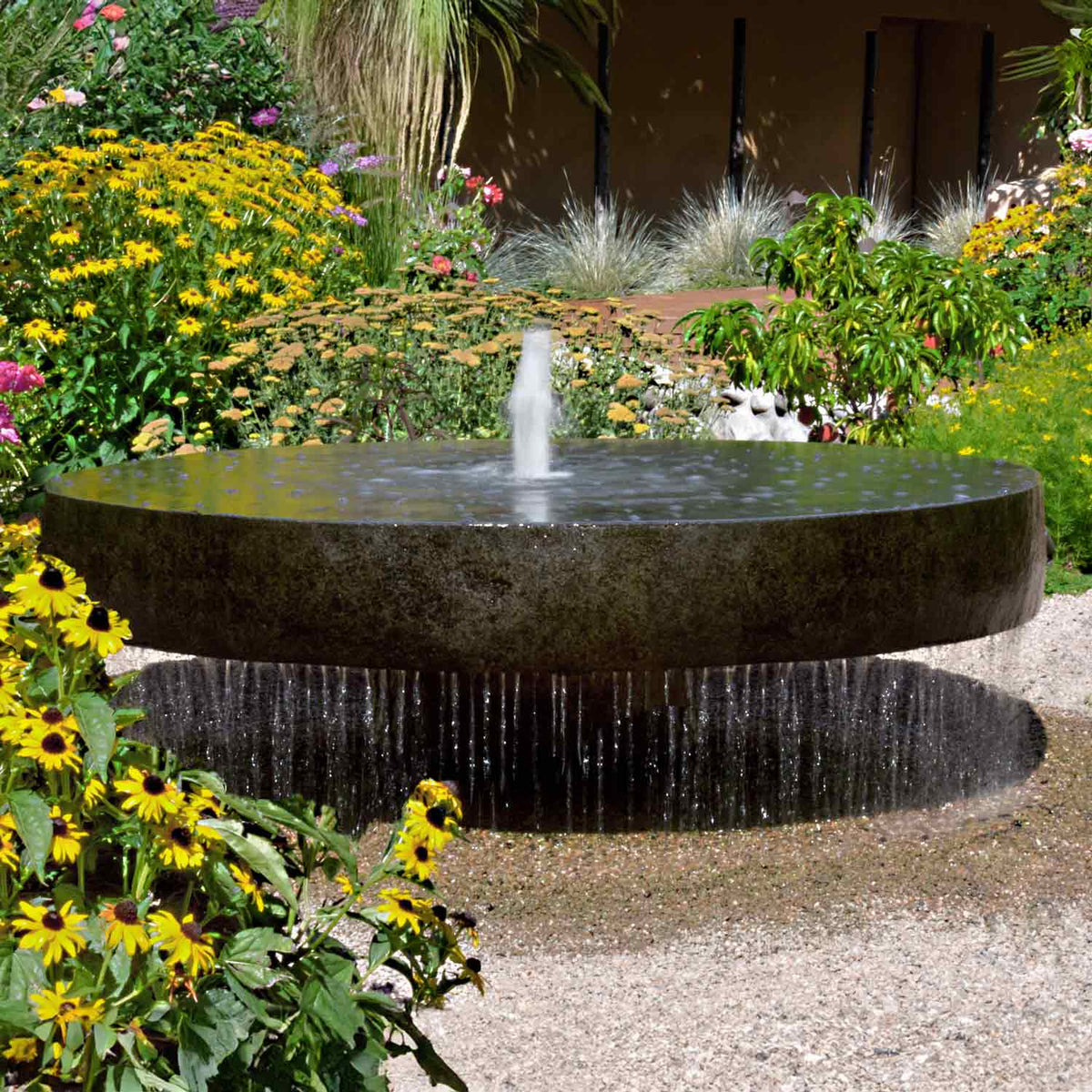 A large natural gray granite millstone fountain with water cascading over the edges, set in a garden with yellow flowers in the foreground. image 1 of 2
