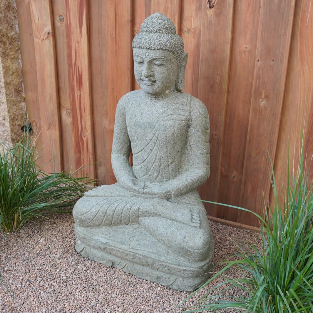 A hand-carved stone Buddha statue, sitting in a meditative pose, made from Verde Basalt, placed on a gravel surface with wooden fence in the background. image 2 of 5