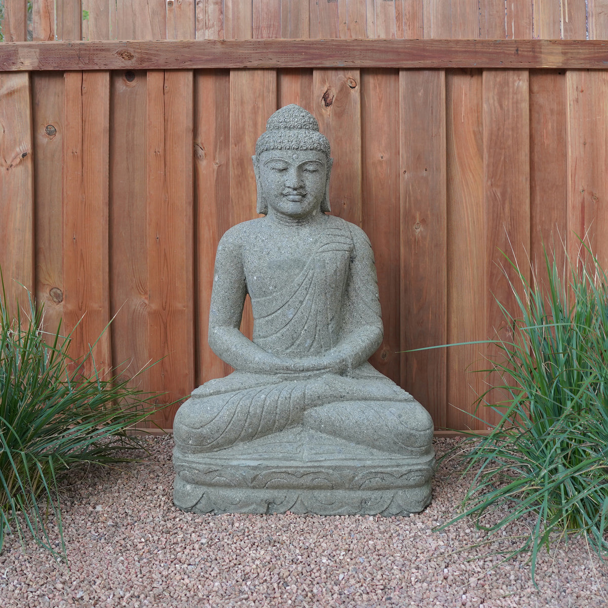 A hand-carved stone Buddha statue, sitting in a meditative pose, made from Verde Basalt, placed on a gravel surface with wooden fence in the background. image 1 of 5