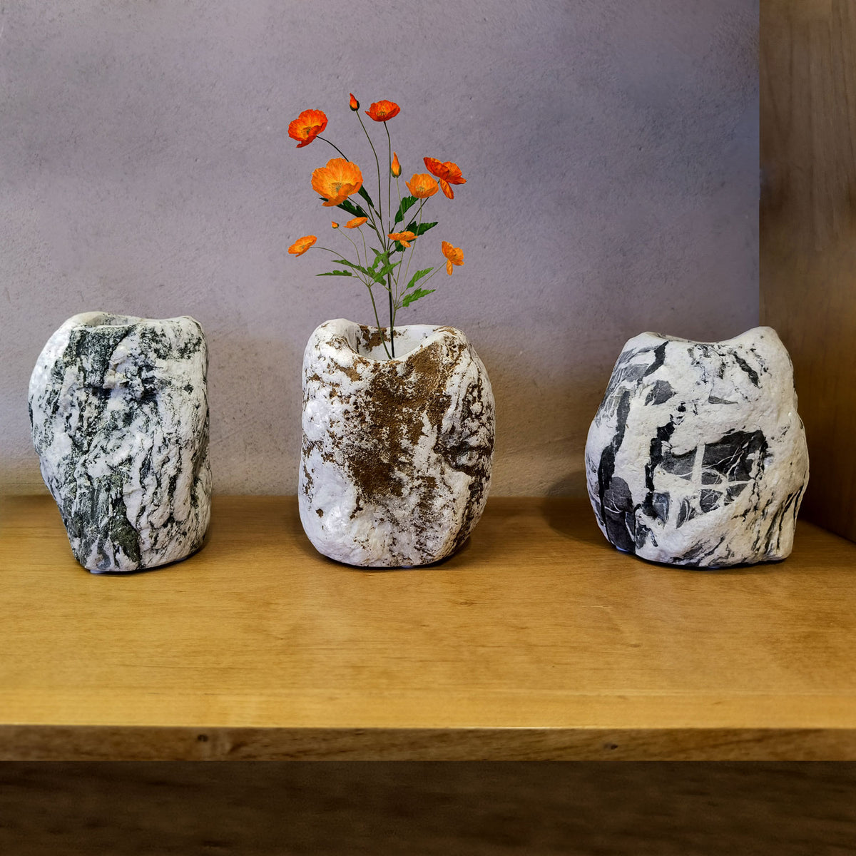 Three natural marble vases in varying shades of gray and one with a chestnut color, each containing a small orange flower, displayed on a wooden shelf. image 1 of 2