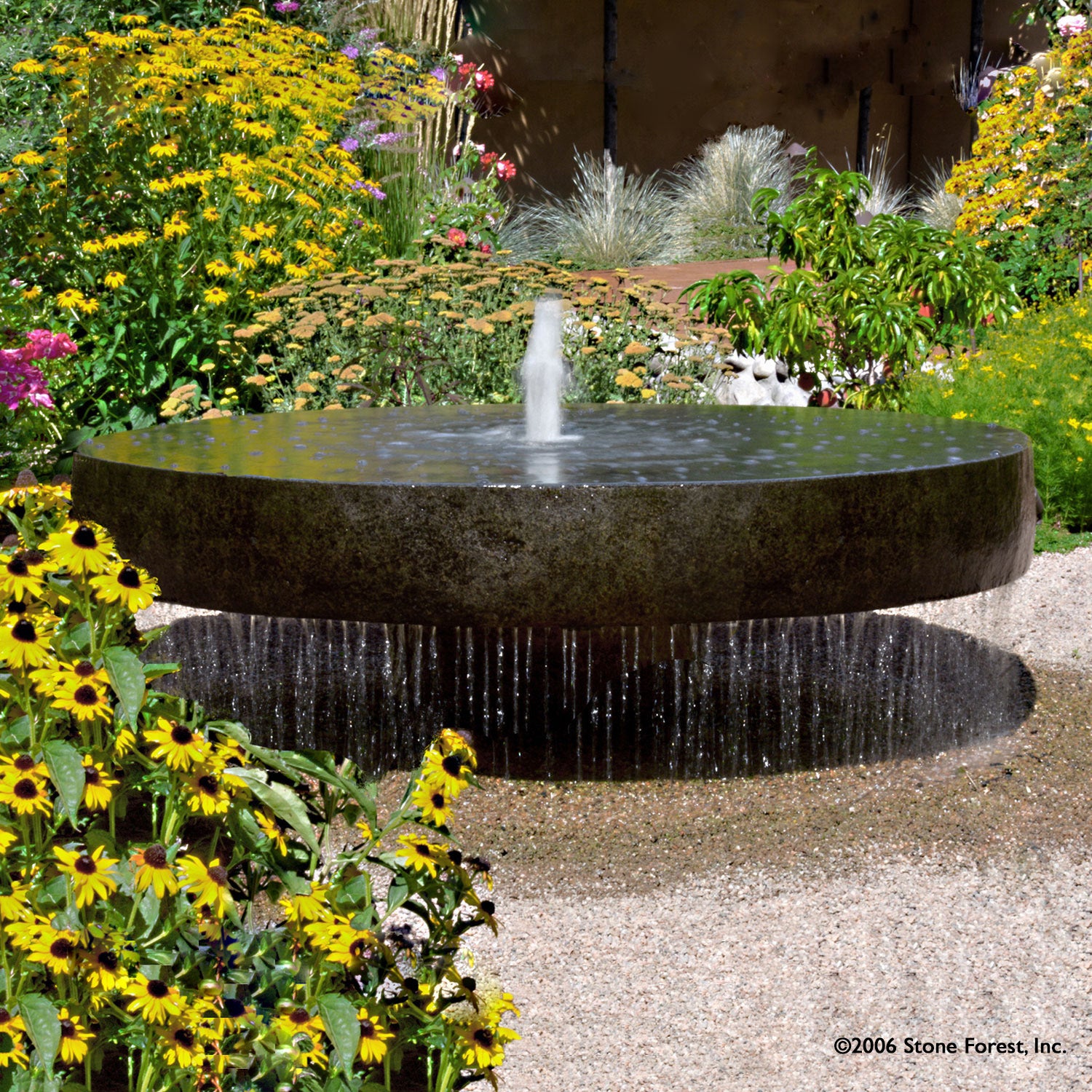 A large natural gray granite millstone fountain with water cascading over the edges, set in a garden with yellow flowers in the foreground.