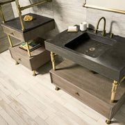 A Terra Bath Sink with aged brass legs, placed on a console stand, accompanied by a drawer vanity with a cement gray wood drawer. The backdrop features a tiled wall and a mirror.