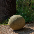 20" Round stone sphere on a gravel surface with a tree and plants in the background