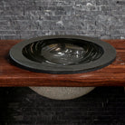 A black granite self-rimming sink placed on an iron stand, with a polished finish and bushed hammer finish on the exterior.
