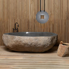 A large, irregularly shaped natural boulder bathtub placed in a bathroom setting with a wood panel wall and a hanging circular decoration above it. The bathtub has a smooth, dark interior and a rough, textured exterior.