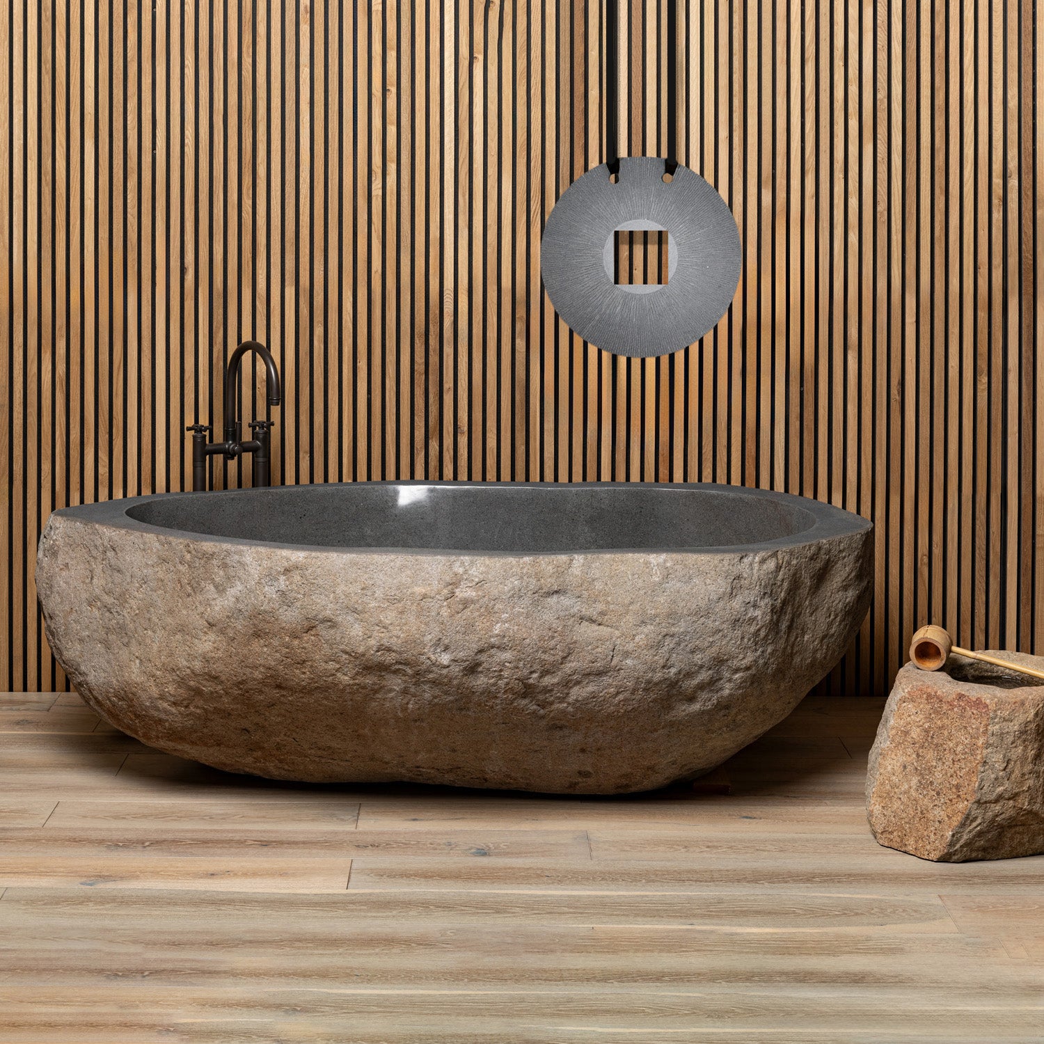 A large, irregularly shaped natural boulder bathtub placed in a bathroom setting with a wood panel wall and a hanging circular decoration above it. The bathtub has a smooth, dark interior and a rough, textured exterior.