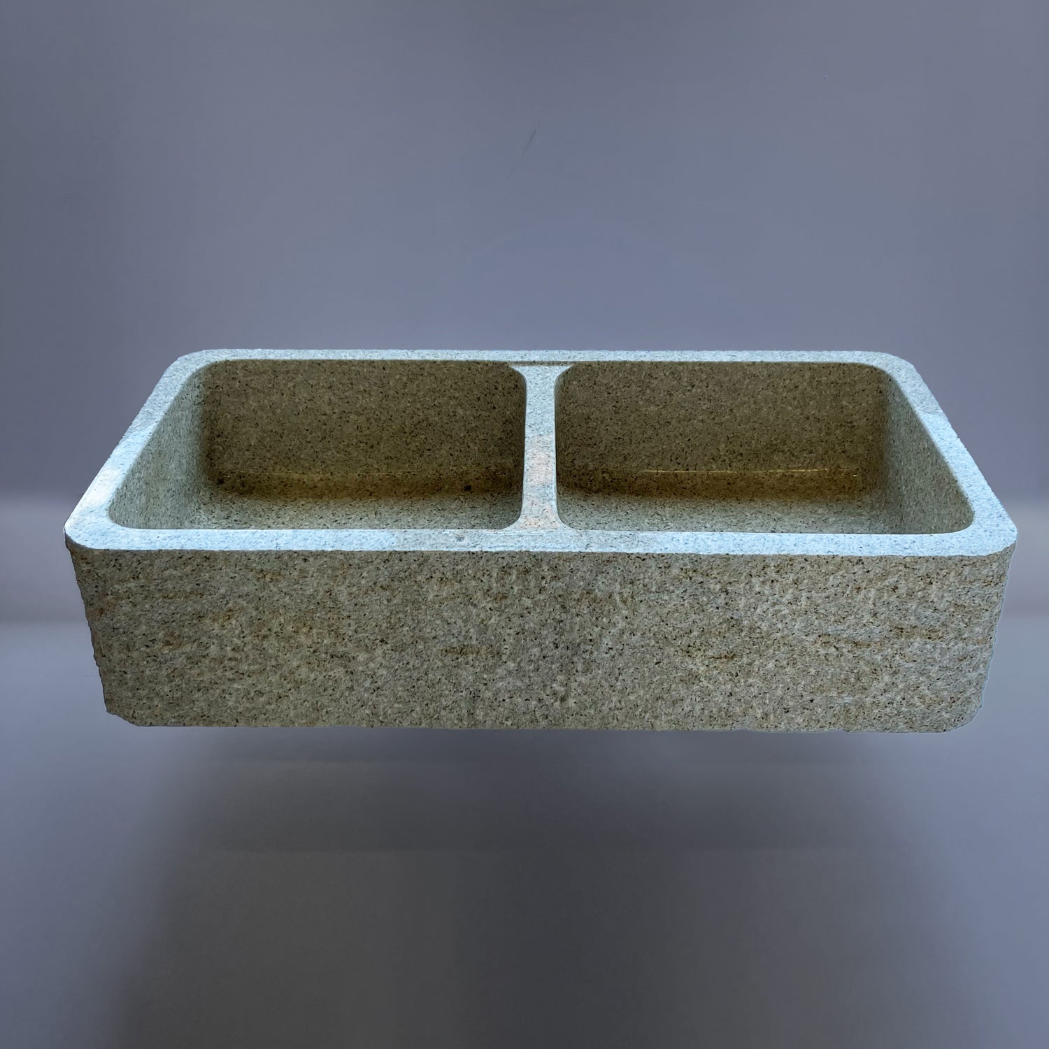 A beige granite double basin farmhouse kitchen sink with a polished interior and a bush hammered apron.