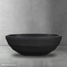 A sleek, monolithic bathtub carved from gray limestone, situated against a dark tiled wall with a dark faucet above it.