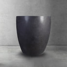 A dark gray, honed basalt soaking tub with a smooth finish and rounded contours, placed on a light gray floor against a textured wall.