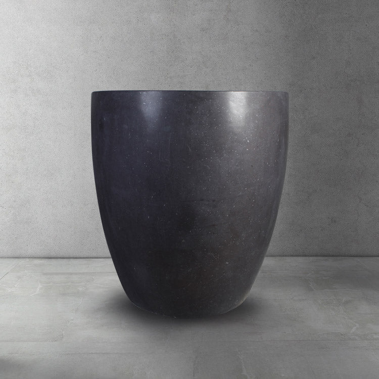 A dark gray, honed basalt soaking tub with a smooth finish and rounded contours, placed on a light gray floor against a textured wall.