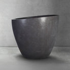 A dark gray, honed basalt soaking tub with a smooth finish and rounded contours, placed on a light gray floor against a textured wall.
