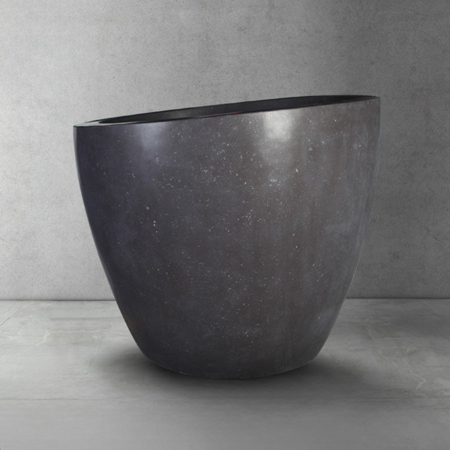 A dark gray, honed basalt soaking tub with a smooth finish and rounded contours, placed on a light gray floor against a textured wall.
