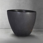 Side view of dark gray, honed basalt soaking tub with a smooth finish and rounded contours, placed on a light gray floor against a textured wall.
