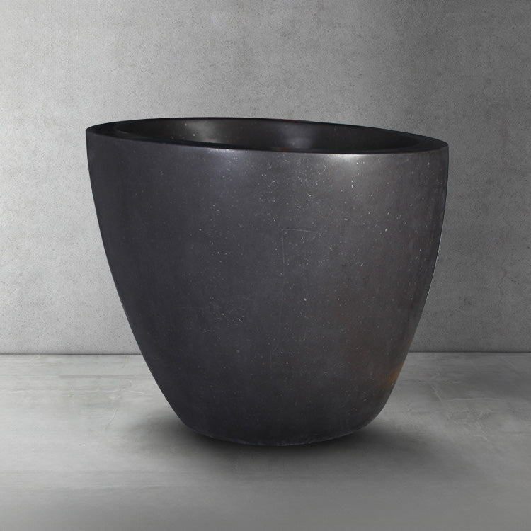 Side view of dark gray, honed basalt soaking tub with a smooth finish and rounded contours, placed on a light gray floor against a textured wall.
