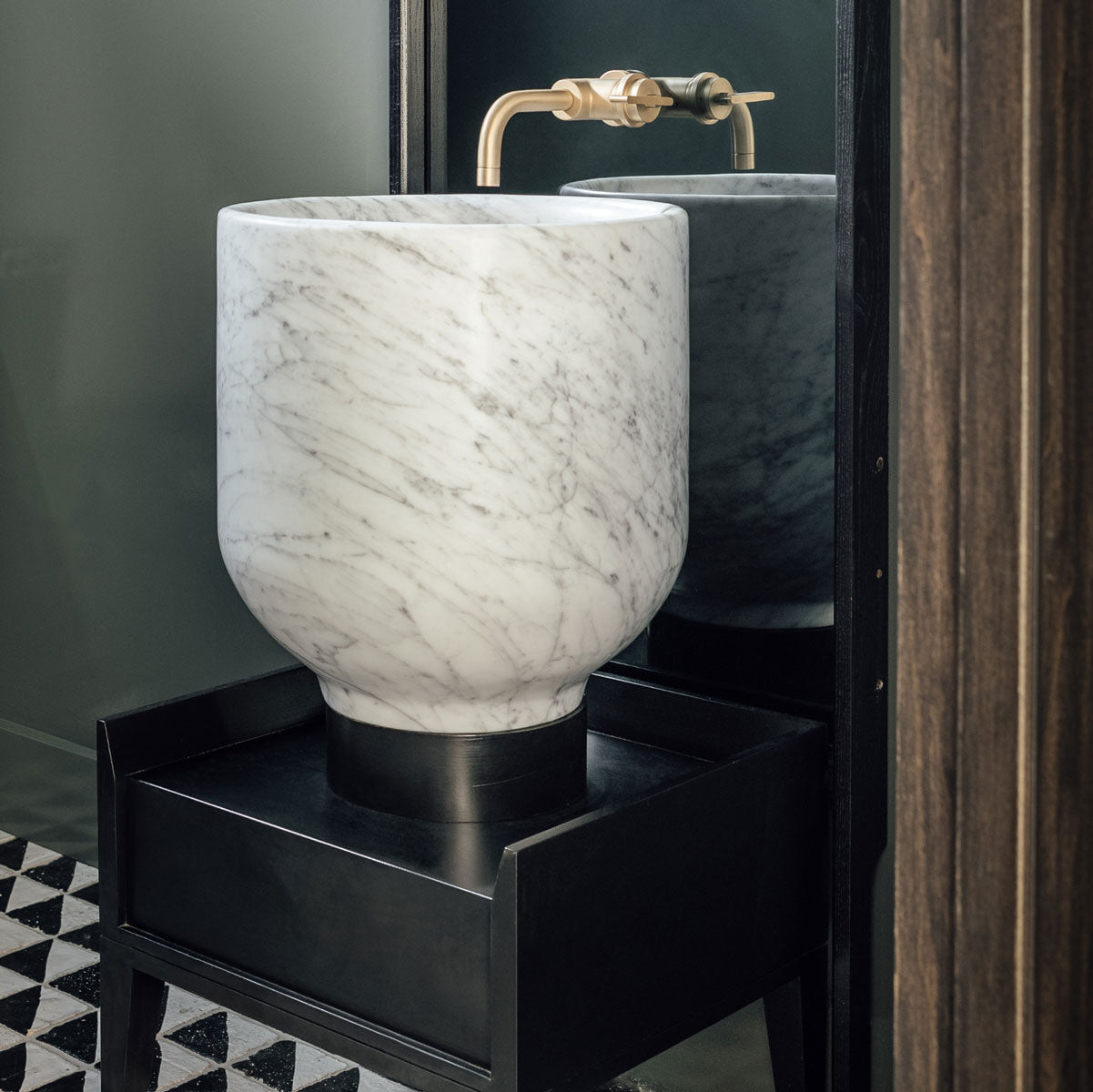 A round white marble vessel sink with gray veining, mounted on a black pedestal table. A brass faucet is visible behind it, and a dark mirror reflects the sink.
