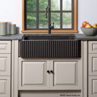 A fluted front farmhouse sink made of noce basalt, installed in a kitchen setting with a black faucet and light-colored cabinetry.