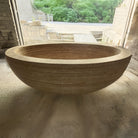 An oval-shaped, beige travertine marble tub positioned indoors with a window in the background.