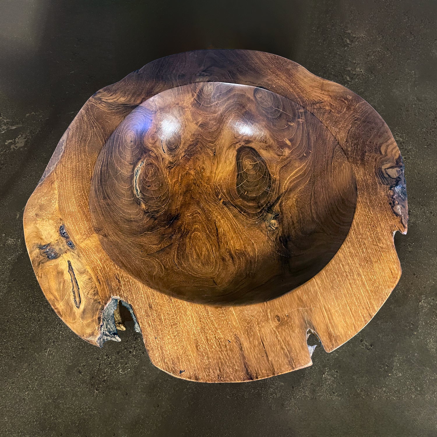 A hand-carved teak wood bowl with a smooth finish, showing natural wood grain, is displayed on a concrete surface 