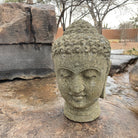 A stone sculpture of a Buddha head with a serene expression, set against a natural outdoor backdrop with rocks and a flowing water feature.