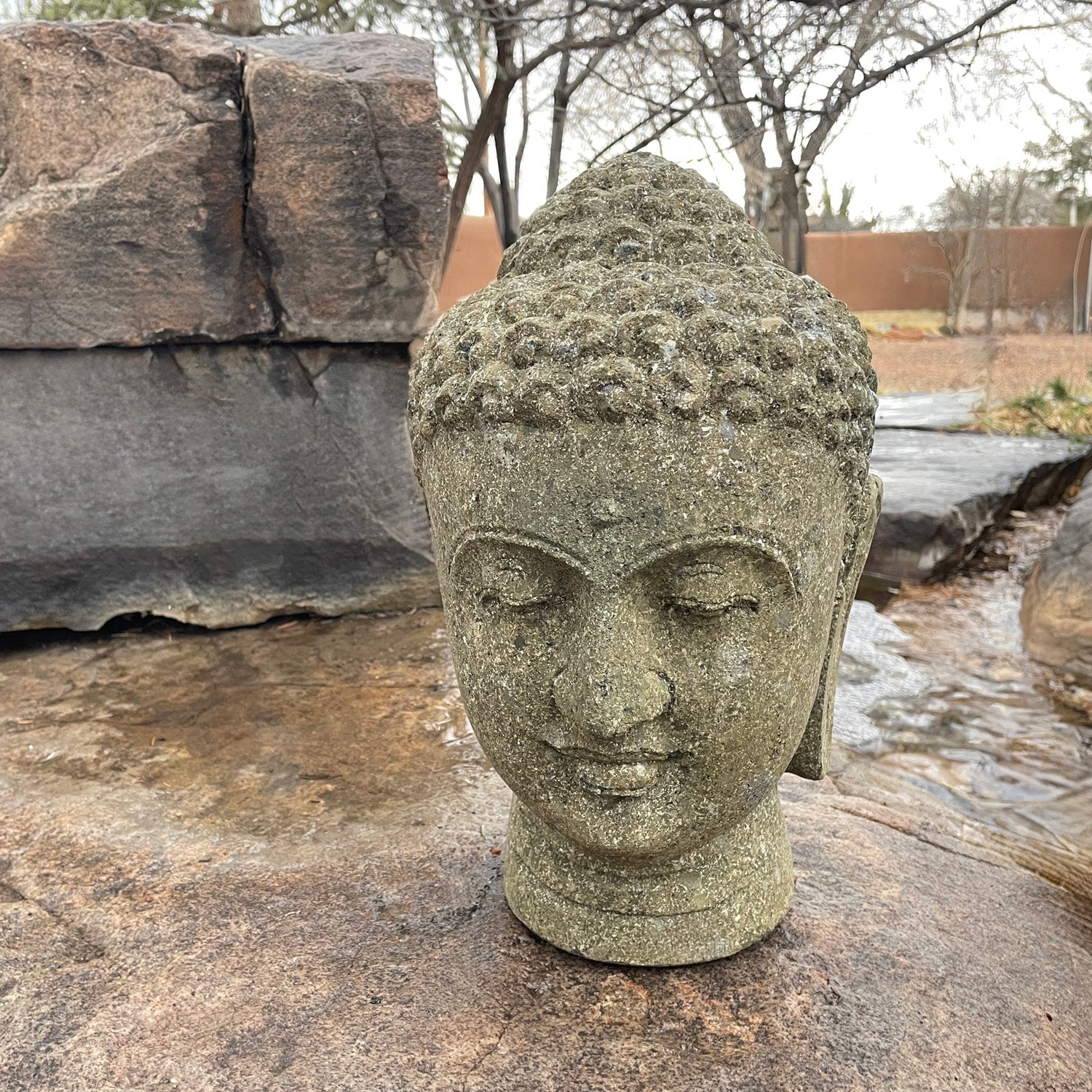 A stone sculpture of a Buddha head with a serene expression, set against a natural outdoor backdrop with rocks and a flowing water feature.