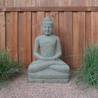 A hand-carved stone Buddha statue, sitting in a meditative pose, made from Verde Basalt, placed on a gravel surface with wooden fence in the background.