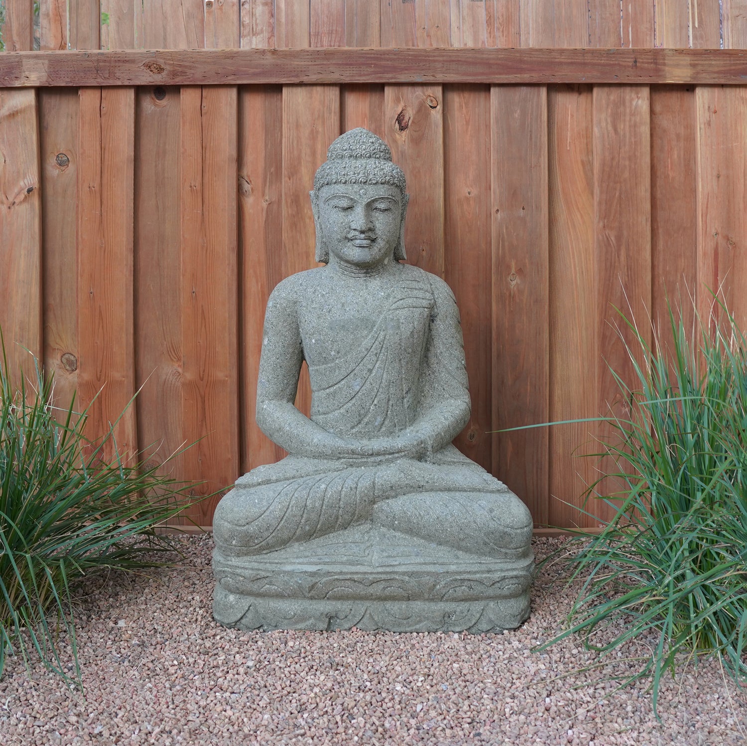 A hand-carved stone Buddha statue, sitting in a meditative pose, made from Verde Basalt, placed on a gravel surface with wooden fence in the background.