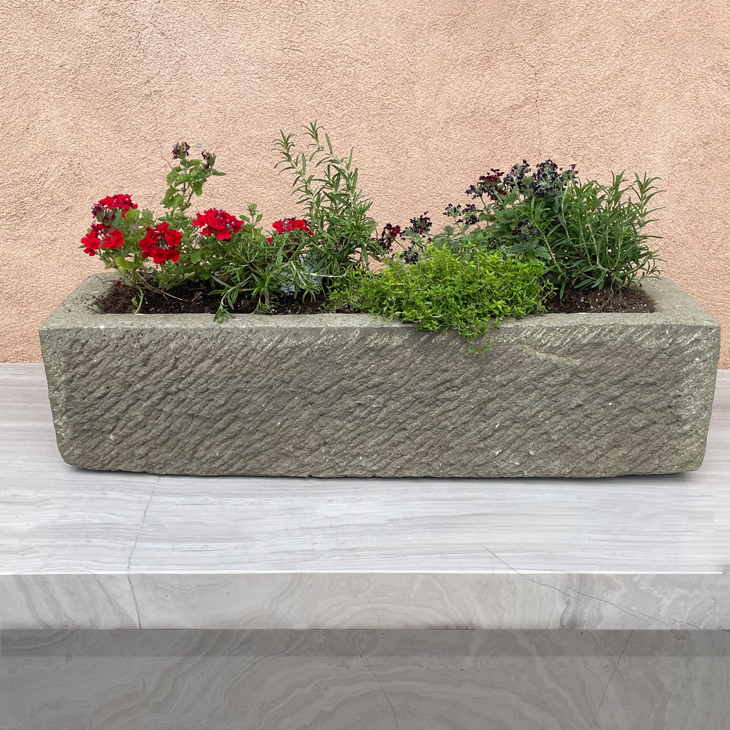Rectangular stone planter with plants on a marble surface against a beige wall.