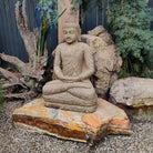 Stone Buddha statue on a rock in a garden setting with rocks and plants.