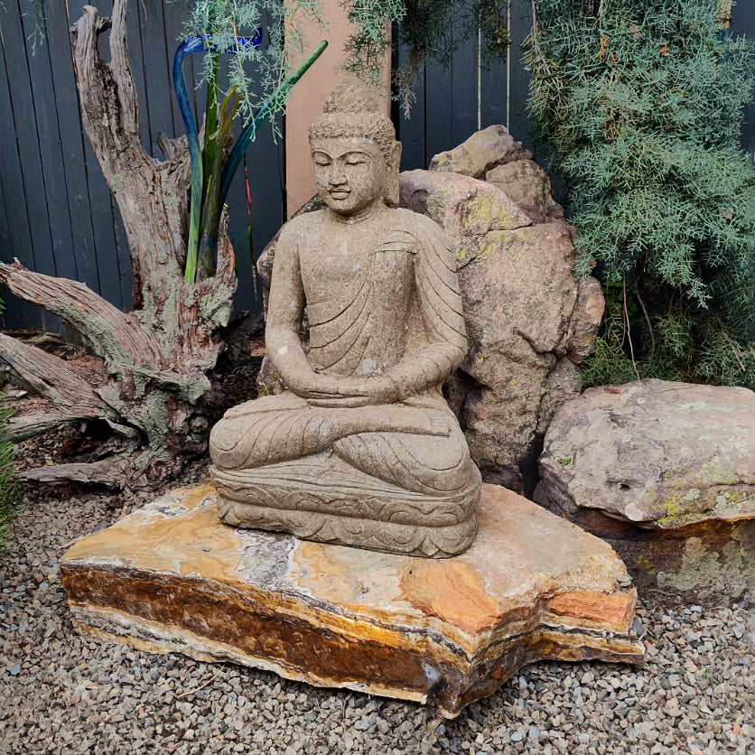 Stone Buddha statue on a rock in a garden setting with rocks and plants.
