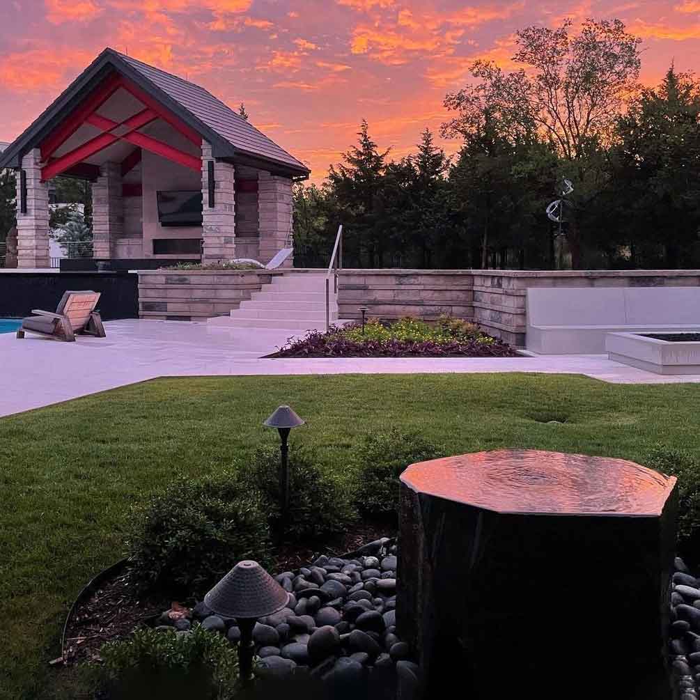A custom fountain with a contemporary design, made of stone, situated in an outdoor setting with a house and garden in the background.