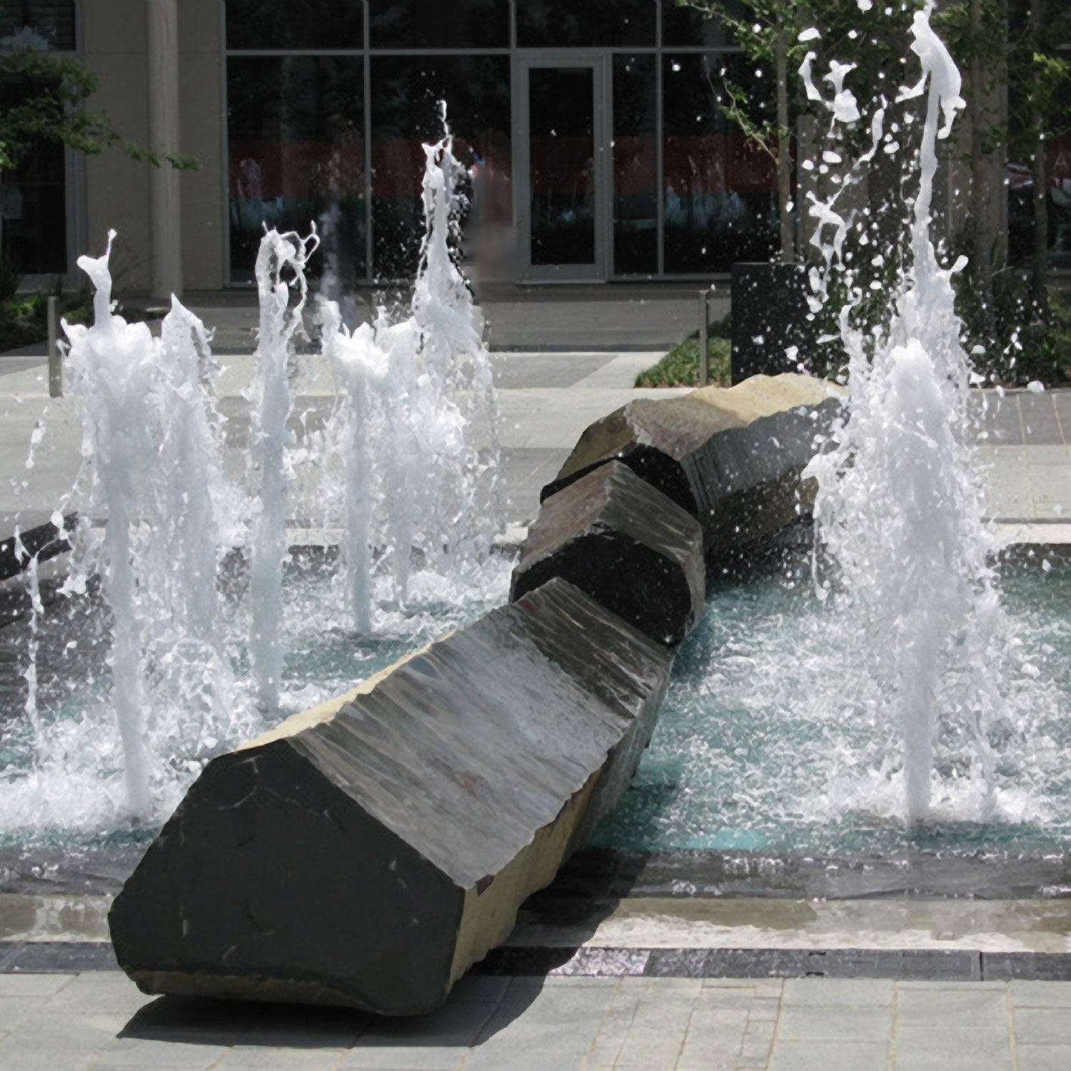 A section of a city park with a large fountain featuring a custom basalt column on which water is spraying, with a blurred background.