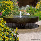 A large natural gray granite millstone fountain with water cascading over the edges, set in a garden with yellow flowers in the foreground.