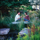Two people sitting on a stone bridge in a garden setting with greenery and a water feature.