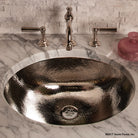 An oval stainless steel sink with a polished finish, with a hammered texture, accompanied by a marble-topped console.