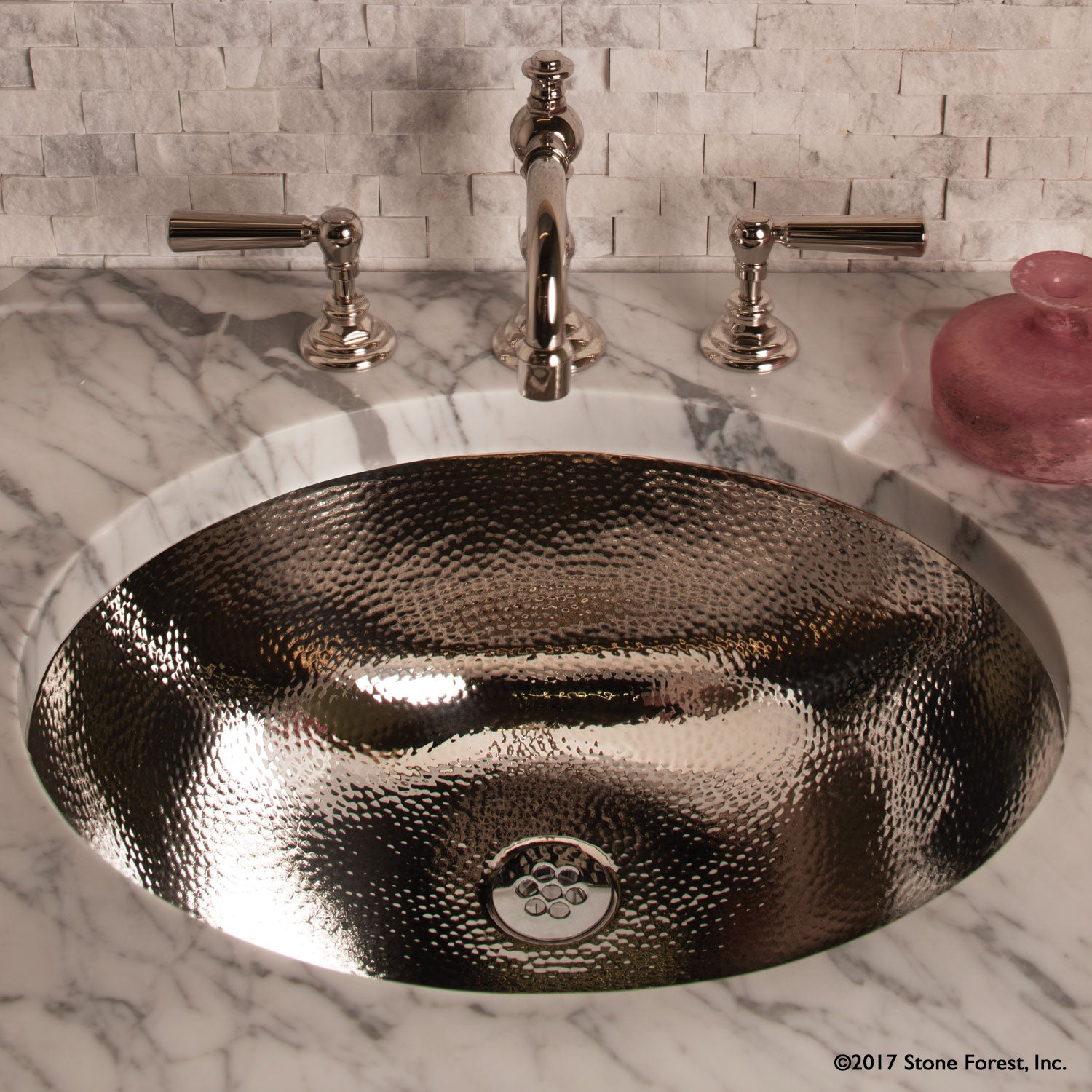 An oval stainless steel sink with a polished finish, with a hammered texture, accompanied by a marble-topped console.